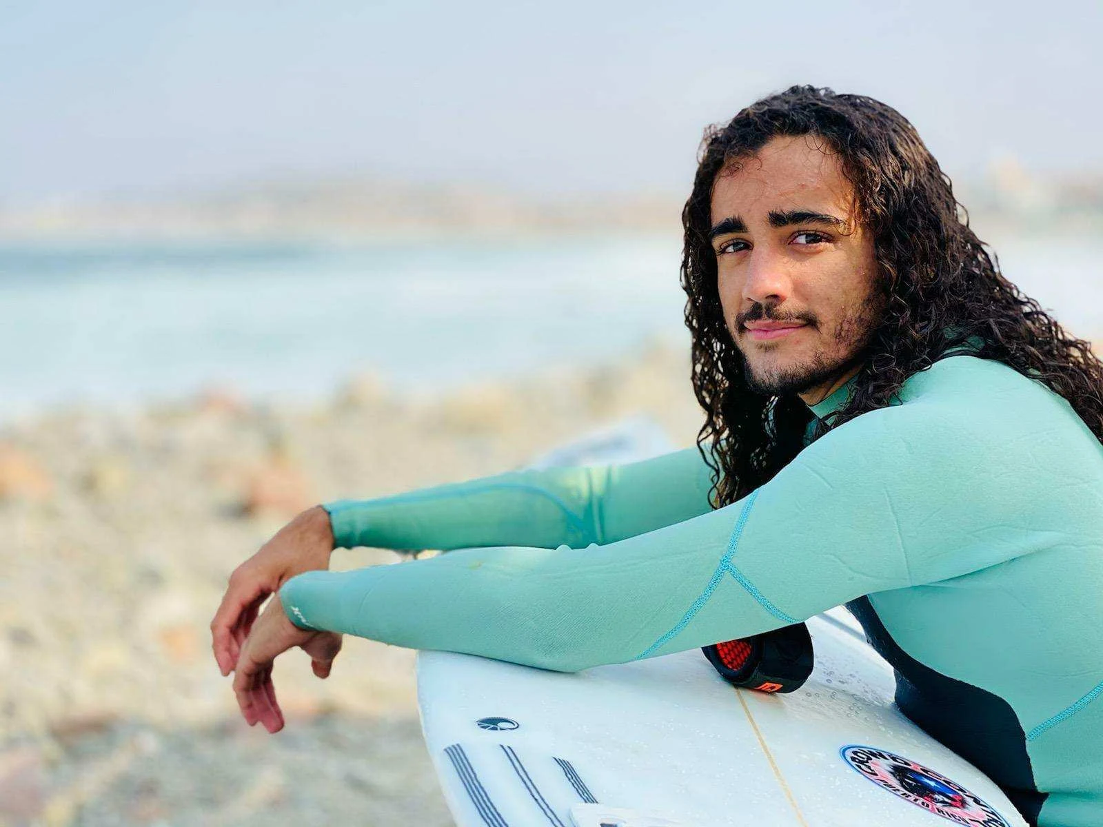 Sabian Cardona Martinez in a turquoise wetsuit resting his arms on a surfboard at the beach